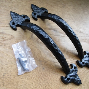 Puede incluir: Dos tiradores de puerta de metal negro con un diseño de flor de lis. Los tiradores son texturizados y tienen un acabado martillado. Se muestran sobre una superficie de madera con una pequeña bolsa de tornillos.