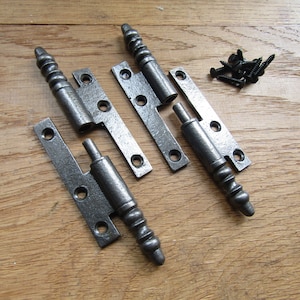 May include: Two pairs of antique-style, black iron hinges with decorative, twisted finials. The hinges are laid out on a wooden surface with a pile of screws.