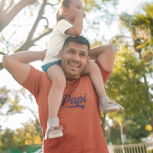 May include: A man wearing an orange t-shirt with the word "Papi" in a yellow diamond shape. He is carrying a young girl on his shoulders. They are both smiling and looking up.