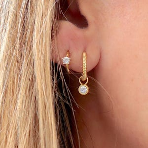 Può includere: Un paio di orecchini a cerchio dorati con un piccolo ciondolo di diamanti. Gli orecchini sono indossati nell'orecchio di una persona con i capelli biondi.