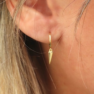 Peut inclure: Gros plan sur une boucle d'oreille dorée. La boucle d'oreille est un petit cercle avec un charm pointu pendant. La boucle d'oreille est suspendue à une oreille. Les cheveux de la personne sont blonds et séparés sur le côté.