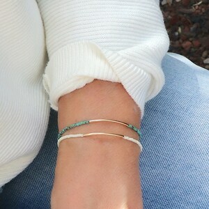 Könnte beinhalten: Zwei Armbänder am Handgelenk einer Person. Das obere Armband besteht aus kleinen türkisfarbenen Perlen und einem dünnen goldenen Band. Das untere Armband besteht aus weißen Perlen und einem dünnen goldenen Band.