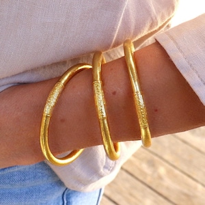 Op de afbeelding: Drie gouden armbanden met ingewikkelde ontwerpen gestapeld op de pols van een persoon.