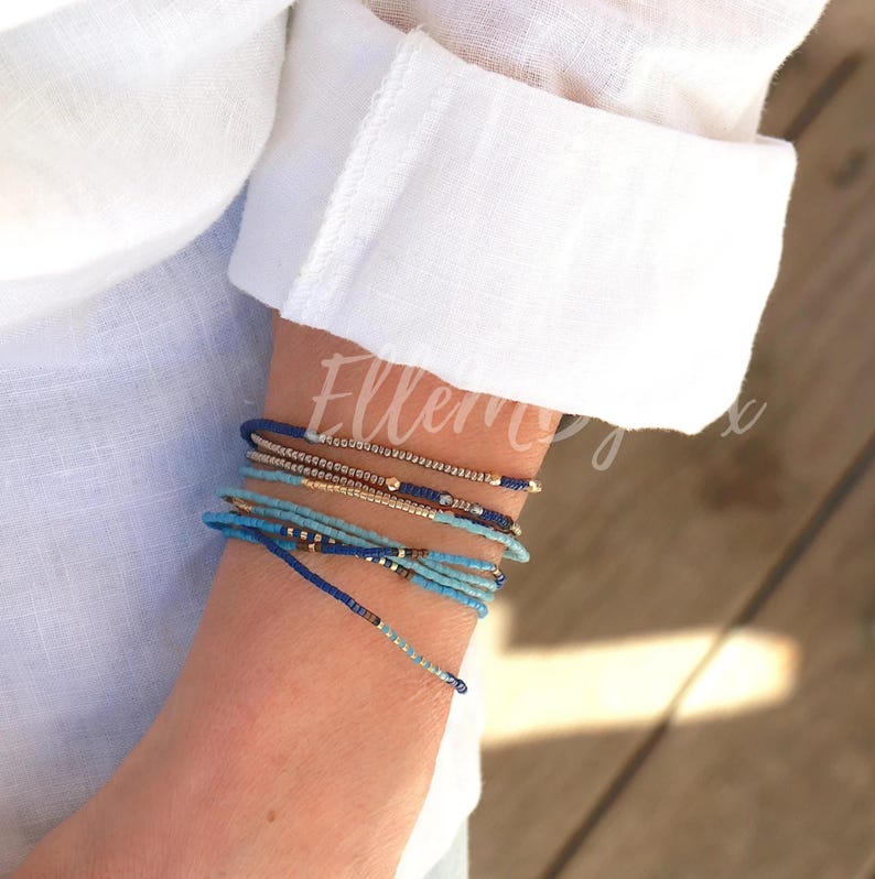 May include: A stack of blue, turquoise, and gold beaded bracelets on a person's wrist. The bracelets are layered and have a delicate, bohemian style.