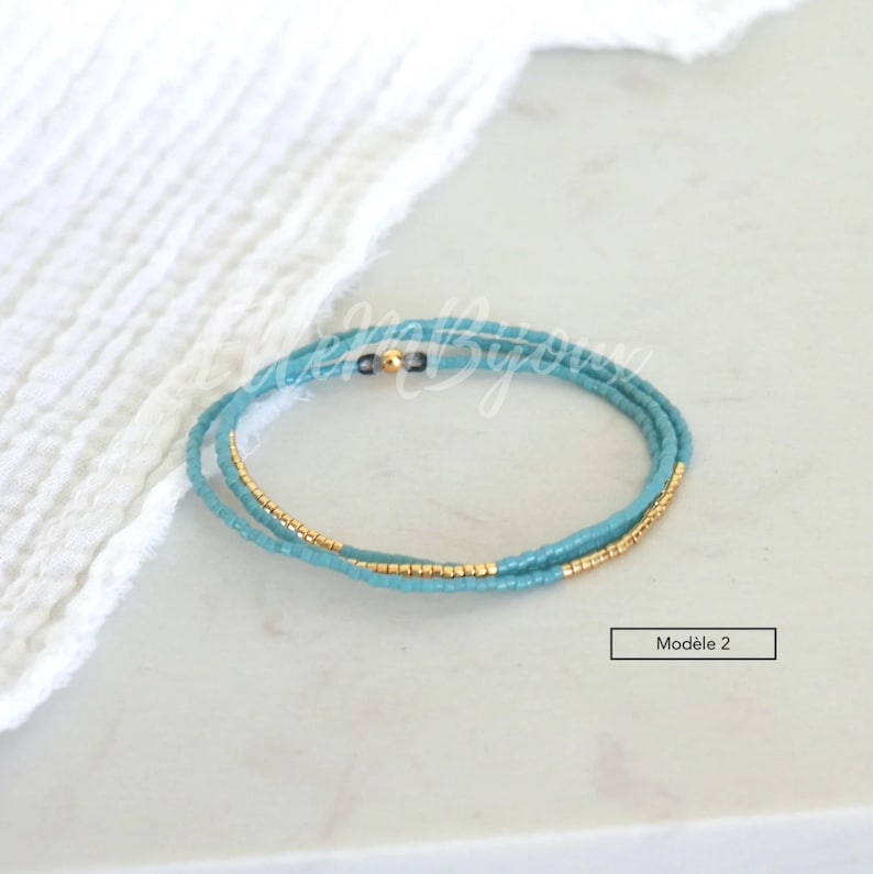 May include: A turquoise and gold beaded bracelet with a clasp. The bracelet is on a white surface.  Mod&egrave;le 2