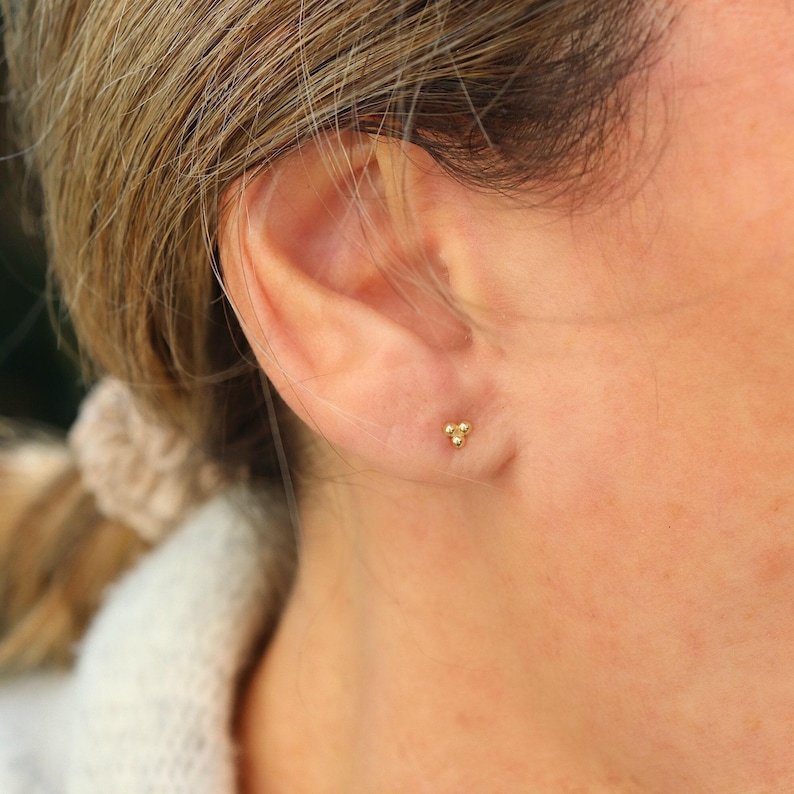 Puede incluir: Primer plano de una oreja con un peque&ntilde;o pendiente de oro con tres peque&ntilde;as bolitas doradas.