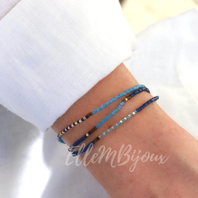 May include: Three layered beaded bracelets in shades of blue with gold accents. The bracelets are stacked on a person's wrist.