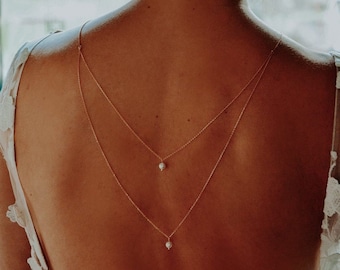 May include: A delicate, layered gold necklace with small, white pearls. The necklace drapes down the back, creating a V-shape. The image shows the back of a person wearing a white lace dress.
