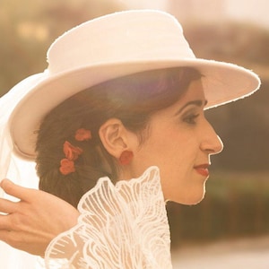 May include: A woman wearing a white lace dress and a white hat with a veil. She has red flowers in her hair and is looking to the right.