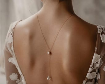 May include: A delicate gold chain necklace featuring a single round pearl accent positioned at the center of the back, with a teardrop-shaped pendant hanging from the end of the chain, worn with a white lace dress with a deep back neckline.