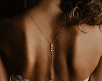 Könnte beinhalten: Eine goldene Kette mit drei kleinen perlmuttartigen Perlen hängt am Rücken einer Person, die ein weißes Spitzenoberteil trägt.