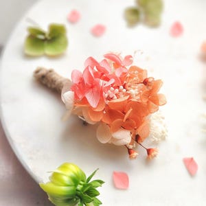 May include: A small, handcrafted floral boutonniere featuring coral and orange dried flowers, accented with white filler flowers. The stem is wrapped in natural twine. Scattered pink petals and green foliage complete the arrangement.
