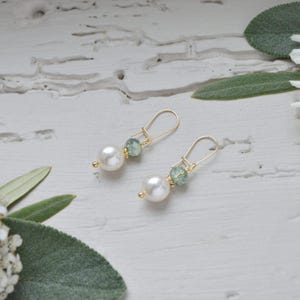 May include: A pair of gold-colored drop earrings. Each earring features a large, white, round pearl and a smaller, light green, faceted bead. The earrings are displayed on a white, textured surface with green foliage.