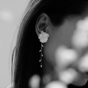 Peut inclure: Gros plan sur l'oreille d'une personne avec une délicate boucle d'oreille en forme de fleur. La boucle d'oreille est en tissu blanc et a une chaîne argentée avec de petites perles qui pendent.