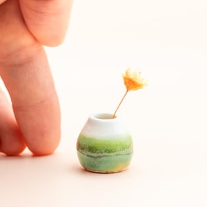 May include: A miniature ceramic vase with a rounded body and a narrow neck. The vase features a white upper section that transitions into horizontal bands of light green and dark green glaze. A single small, dried yellow flower sits inside.