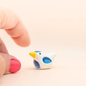 May include: A small ceramic bird figurine with blue and yellow accents. The bird is white with blue wings and a yellow beak. The figurine is sitting on a light pink surface.