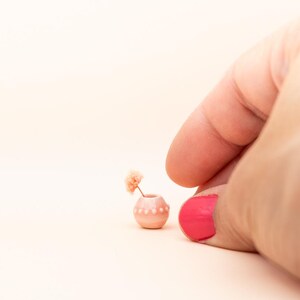 May include: A tiny pink ceramic vase, decorated with white dots, contains a single peach-coloured flower. The miniature vase is designed for decorative purposes. The background is a soft, peachy hue.