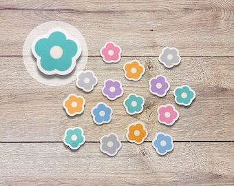 May include: Assortment of colorful flower-shaped stickers in various shades of blue, pink, orange, and gray. The stickers are arranged in a semi-circle on a light brown wooden surface.