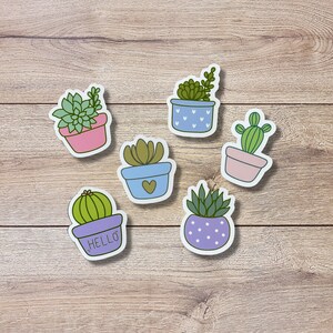 May include: Six colorful succulent plant stickers with different designs. The stickers are on a light brown wooden background. The stickers feature different types of succulents in pots with various colors and patterns. One sticker has the word "HELLO" on it.