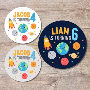 May include: Two round, colorful birthday party stickers with a space theme. One sticker is white with a yellow rocket, blue earth, and orange, red, and gray planets. The text reads "Jacob is turning 4". The other sticker is dark blue with a yellow rocket, blue earth, and orange, red, and gray planets. The text reads "Liam is turning 6".