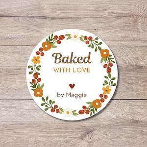 May include: A round sticker with the words "Baked WITH LOVE" in brown font, surrounded by a floral wreath of orange flowers, green leaves, and red berries. The name "by Maggie" is below the heart symbol.
