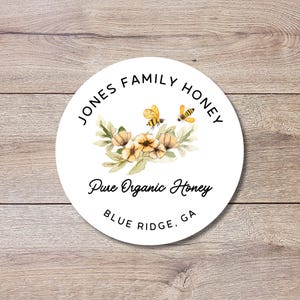 May include: A round white label with the text "JONES FAMILY HONEY" in a circular arrangement. The label features a watercolor illustration of bees and flowers, with the words "Pure Organic Honey" and "BLUE RIDGE, GA" below.