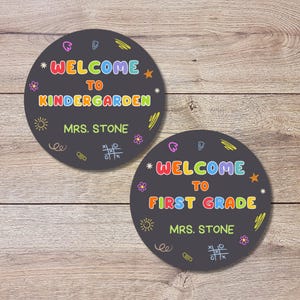 May include: Two round, dark gray signs with colorful text and doodles. One says "WELCOME TO KINDERGARTEN MRS. STONE." The other says "WELCOME TO FIRST GRADE MRS. STONE."