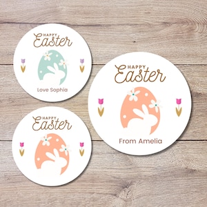 Puede incluir: Tres pegatinas redondas blancas con un diseño de huevo de Pascua rosa pastel y azul. Las pegatinas presentan una silueta de conejo dentro del huevo y el texto "Feliz Pascua". Una pegatina dice "Love Sophia" y otra dice "From Amelia".