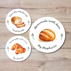 May include: Three round white stickers with black text and images of baked goods. The first sticker says "From the Kitchen of Carol Smith" and has a loaf of white bread with slices. The second sticker says "Homemade with Love By Stephanie" and has a round loaf of bread with sesame seeds. The third sticker says "Homemade with Love By Amelia" and has two croissants.