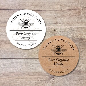 May include: Two round labels for honey products. One is white, the other is brown. Both feature a detailed bee illustration and the text "MADERA HONEY FARM", "Pure Organic Honey", and "BLUE RIDGE, GA". The labels are on a wooden surface.