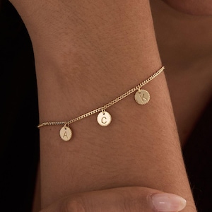 Peut inclure: Un bracelet doré avec une fine chaîne et quatre petits charms ronds. Chaque charm porte une lettre : A, C et K. Le bracelet est porté sur un poignet, mettant en valeur son design épuré.