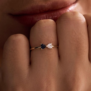 Könnte beinhalten: Ein goldener Ring mit zwei herzförmigen Edelsteinen, einem schwarzen und einem weißen. Der Ring ist filigran und sitzt an einem Finger. Schmuck.