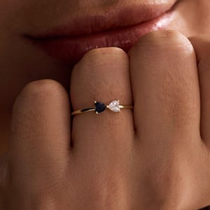 Puede incluir: Un anillo de oro con dos piedras preciosas en forma de corazón, una negra y otra blanca. El anillo se lleva en un dedo, con las piedras colocadas una al lado de la otra. El delicado diseño del anillo se destaca sobre un fondo suave.