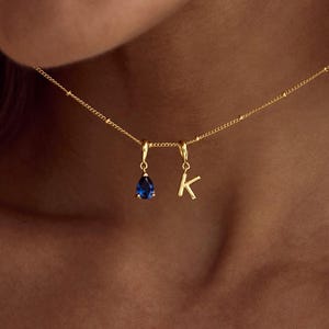 Könnte beinhalten: Eine goldfarbene Halskette mit einem kleinen blauen tropfenförmigen Anhänger und einem Buchstabenanhänger "K". Die Halskette wird um den Hals getragen. Der Hintergrund ist unscharf.