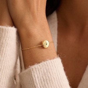 May include: A gold bracelet with a small, round charm featuring a green gemstone. The bracelet is delicate, with a thin chain. The charm is centered on the wrist, creating a simple and elegant look.