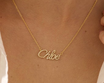 Puede incluir: Un collar dorado con el nombre "Chloe" escrito en cristales claros y brillantes. El collar se muestra sobre una camisa blanca y piel.