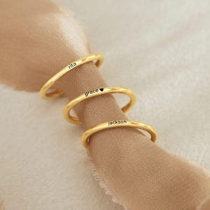 Könnte beinhalten: Drei goldene Ringe mit den eingravierten Namen "Rita", "Grace" und "Jackson". Der mittlere Ring hat ein kleines Herzsymbol neben dem Namen eingraviert.