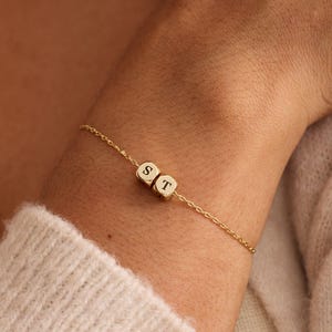 Op de afbeelding: Een delicate gouden kettingarmband met twee kleine, goudkleurige kubusvormige kralen. Elke kubus heeft een enkele letter, "S" en "T", in een eenvoudig, elegant lettertype. De armband wordt om een pols gedragen, wat de gepersonaliseerde sieraden laat zien.