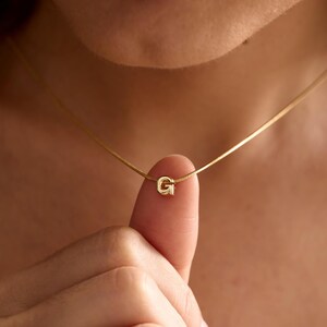 May include: A gold-toned necklace featuring a delicate chain and a small, centred letter G pendant. The pendant is a polished, metallic initial charm suspended on a thin, smooth chain that rests against the neck, showcasing a simple design.
