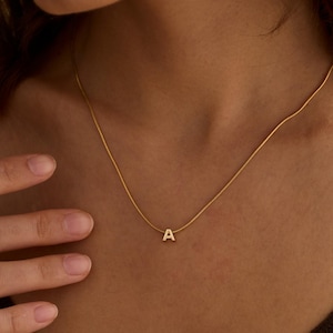May include: A gold-toned necklace featuring a delicate snake chain and a small, centered letter A pendant. The pendant is polished and minimalist in design, resting against the collarbone. The chain is thin and smooth, providing a subtle shine.