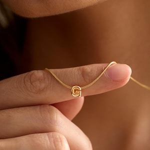 May include: A gold-toned necklace featuring a delicate snake chain and a small, rounded letter G pendant. The pendant is centred on the chain, which rests against the skin. The metal has a polished, reflective finish throughout the piece.