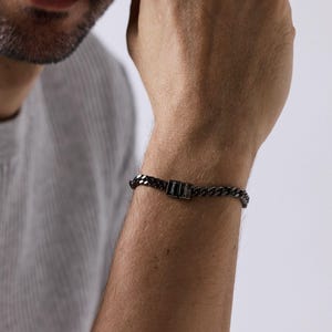 Könnte beinhalten: Ein schwarzes Kettenarmband mit rechteckigem Verschluss. Das Armband hat ein Panzerketten-Design. Der Verschluss ist klein und rechteckig. Das Armband wird am Handgelenk getragen. Das Metall ist dunkel.