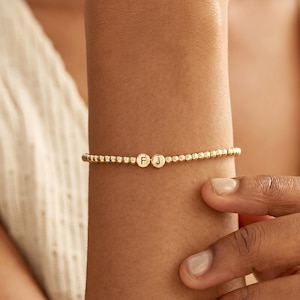 May include: A gold-toned beaded bracelet featuring two small, circular charms positioned in the centre. Each charm is engraved with a single uppercase letter, specifically the letters F and J, displayed in a clear, dark, sans-serif font.