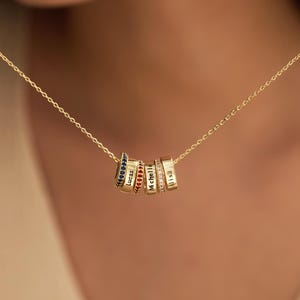 Peut inclure: Un collier à chaîne dorée avec cinq breloques cylindriques. Les breloques sont gravées avec les prénoms Lucas, Michelle et Alisa, et sont ornées de pierres incrustées bleues, rouges et transparentes. Le collier est porté sur une chaîne dorée.