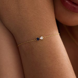 Könnte beinhalten: Ein zartes Armband aus einer goldfarbenen Kette mit zwei kleinen herzförmigen Steinen, einem dunkelblauen und einem klaren, die nebeneinander in der Mitte angeordnet sind. Die feinen Metallglieder verbinden die Steine zu diesem Armschmuck.
