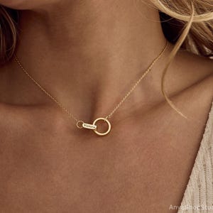 May include: A gold-colored necklace featuring two interlocking circles. One circle is larger and the other is smaller with the name "Victoria" engraved on it. The necklace is displayed on a person's neck.