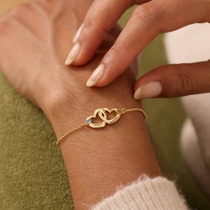 Puede incluir: Una pulsera dorada con dos colgantes entrelazados en forma de corazón. Cada corazón tiene una pequeña piedra preciosa y texto grabado. La pulsera está unida a una fina cadena.