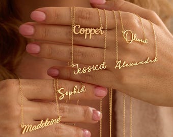 May include: Gold-colored name necklaces spelling out "Copper", "Olivia", "Jessica", "Alexandra", "Sophia", and "Madeleine" in a cursive font. The necklaces are displayed against a blurred background, showcasing the delicate jewelry.