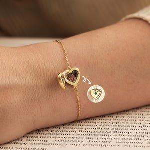 Könnte beinhalten: Ein goldenes Kettenarmband mit einem herzförmigen Medaillon-Anhänger. Das Medaillon ist geöffnet und zeigt eine kleine gepresste rosa Blume im Inneren. Ein kreisförmiger Ausschnitt zeigt das geschlossene Medaillon mit einem kleinen grünen Stein.