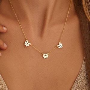 Puede incluir: Collar de oro con tres dijes de huella de pata, cada uno grabado con un nombre: "Daisy", "Eva" y "Charlie".